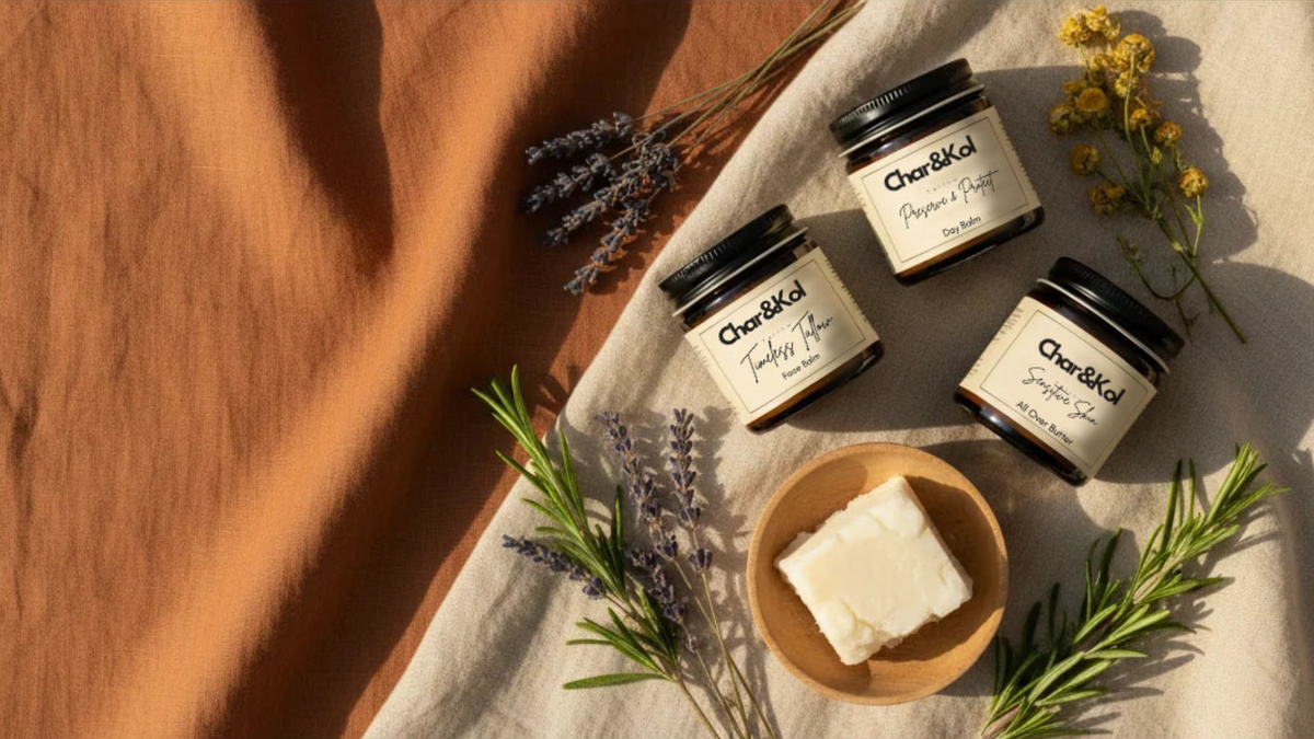 Three jars of Char&Kol Tallow skincare  arranged on a draped fabric surface with dried flowers, rosemary, and a small bowl of white soap. The scene is styled in warm natural light with a terracotta fabric panel on the left.